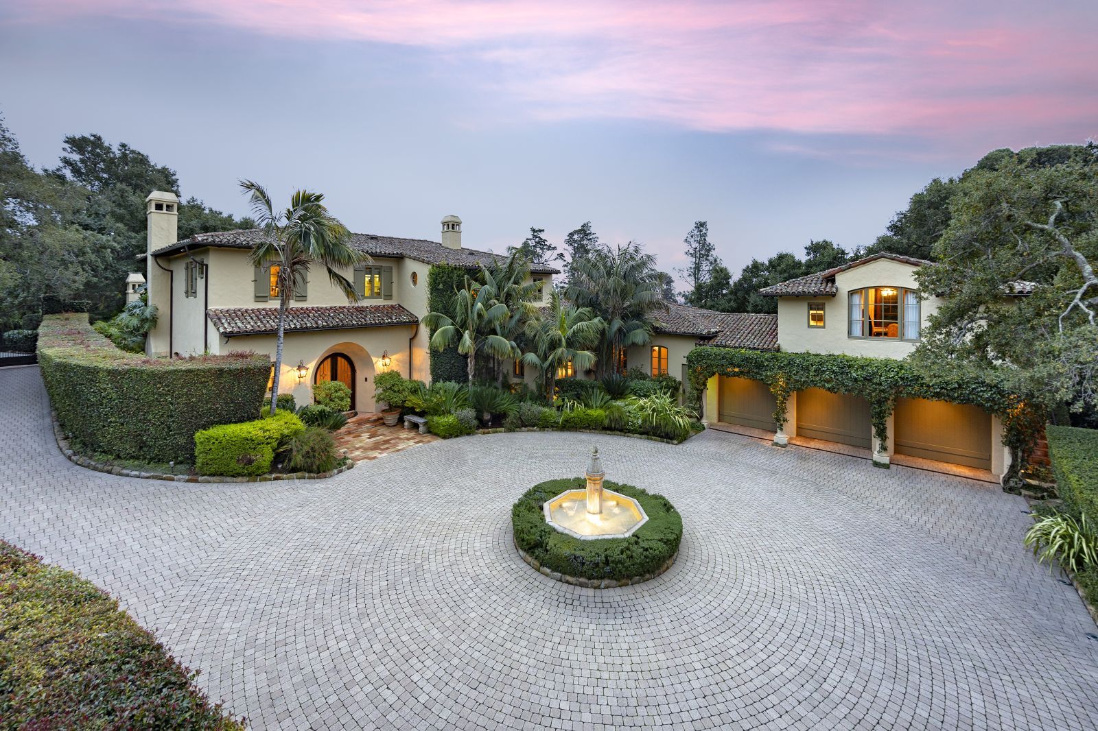 The front of a world-class Mediterranean estate with a brick-paved courtyard encircling a fountain, with the luxury home behind it and a pink and blue sky and lush landscaping.